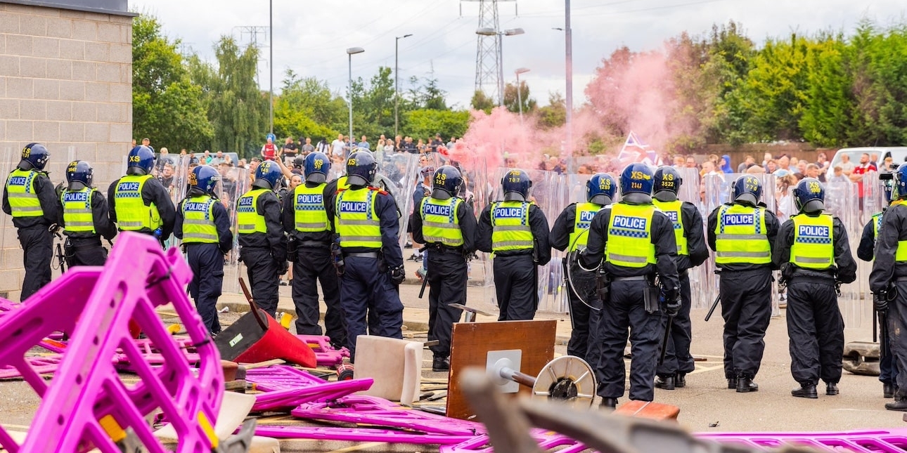 A line of uniformed riot police officers stand in front of damaged furniture and plastic barriers outside a hotel. In the distance are hundreds of protesters, pink smoke and flags